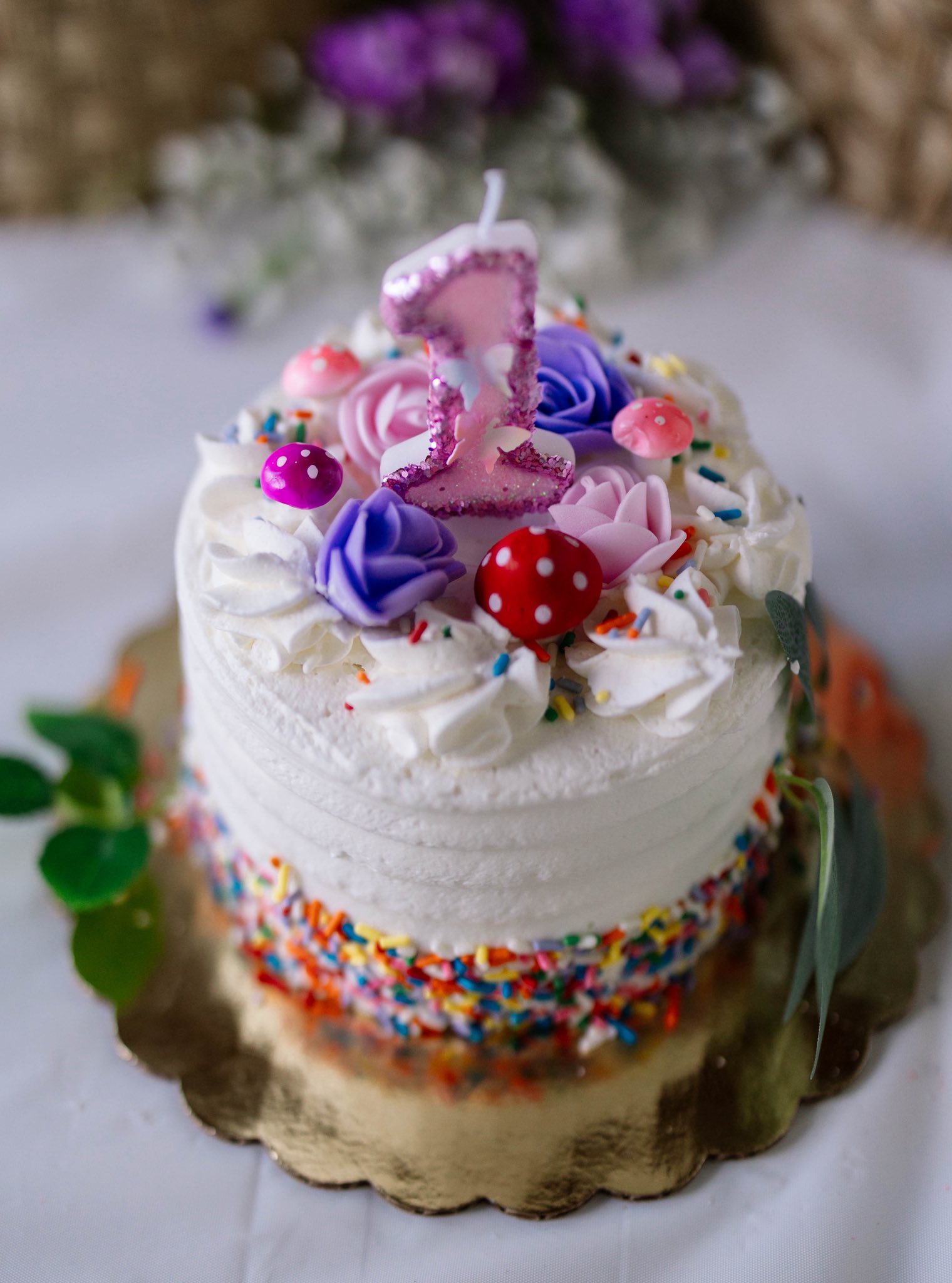 Small round cake with white frosting, colorful sprinkles, sugar flowers, and a pink number one candle