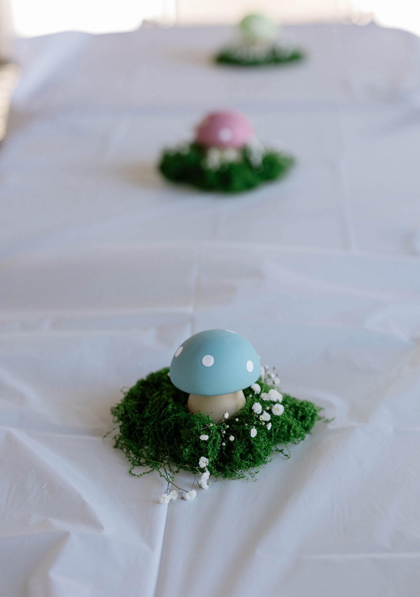 Blue, pink, and green decorative mushrooms in moss and baby's breath floral settings on a white tablecloth.