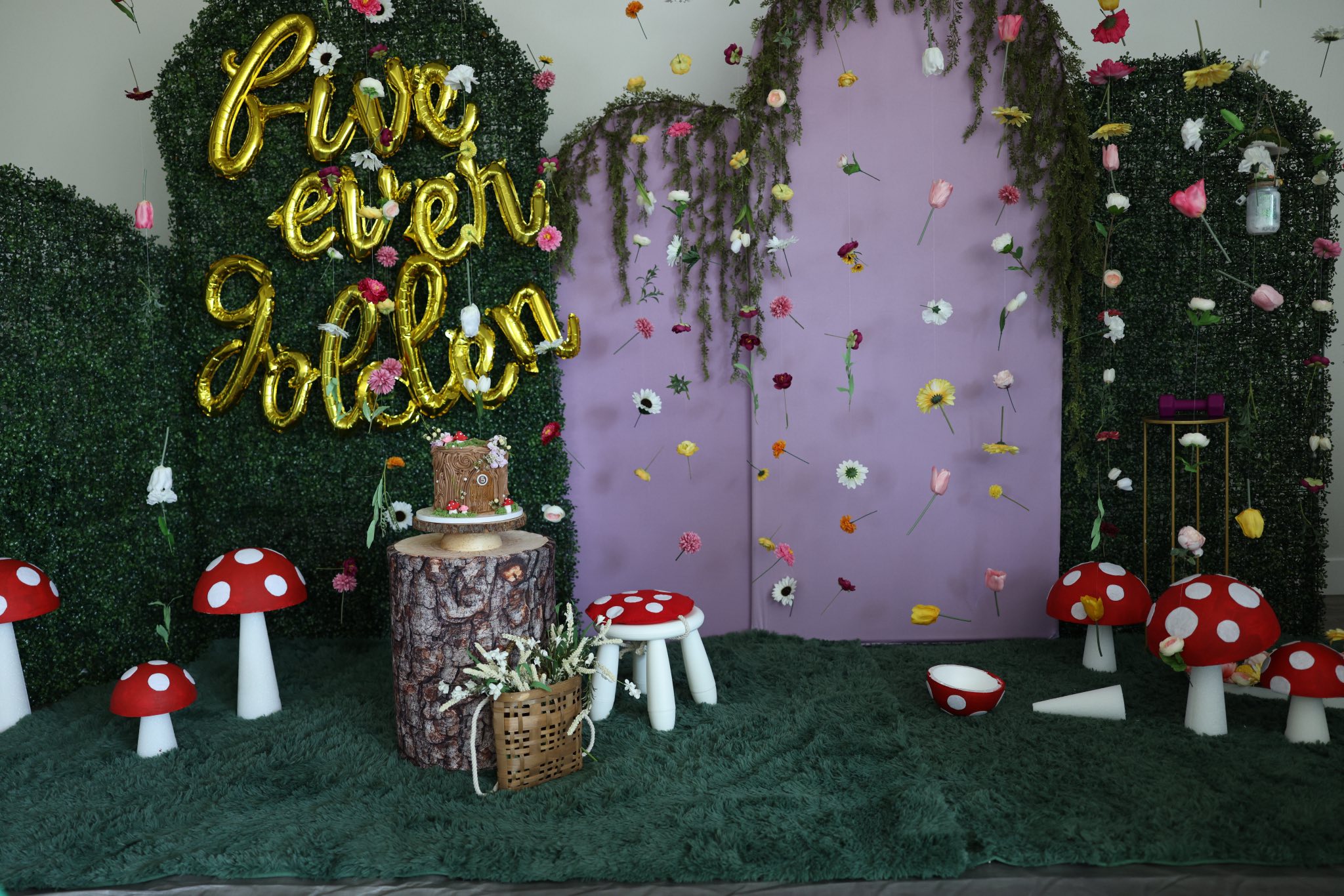 Boho woodland party backdrop featuring mushroom decor, a birthday cake on a stump, and five golden balloon script
