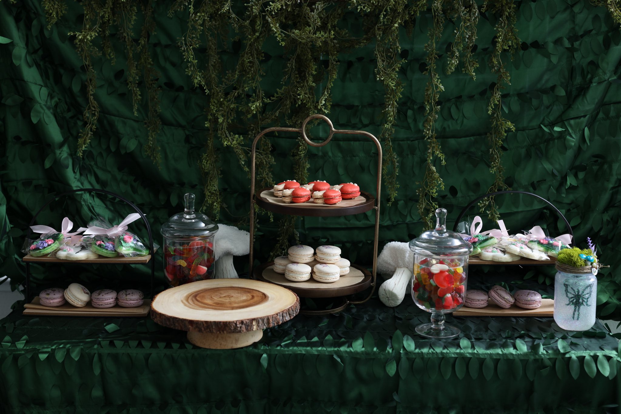 Forest-themed dessert table featuring tiered trays, red and white macarons, a wooden cake stand, and candy jars.