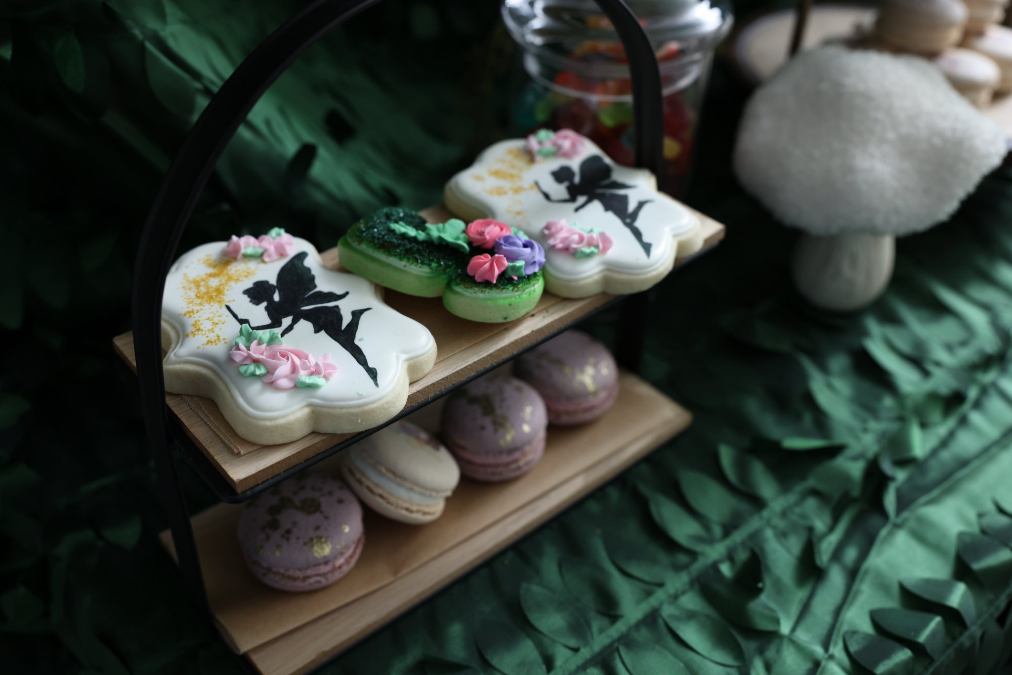 Two-tiered display stand featuring fairy-decorated sugar cookies and assorted colorful macarons.
