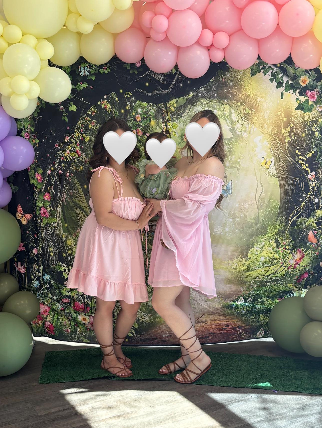 Two women wearing matching pink dresses holding a baby in front of a forest-themed event backdrop with pastel balloons