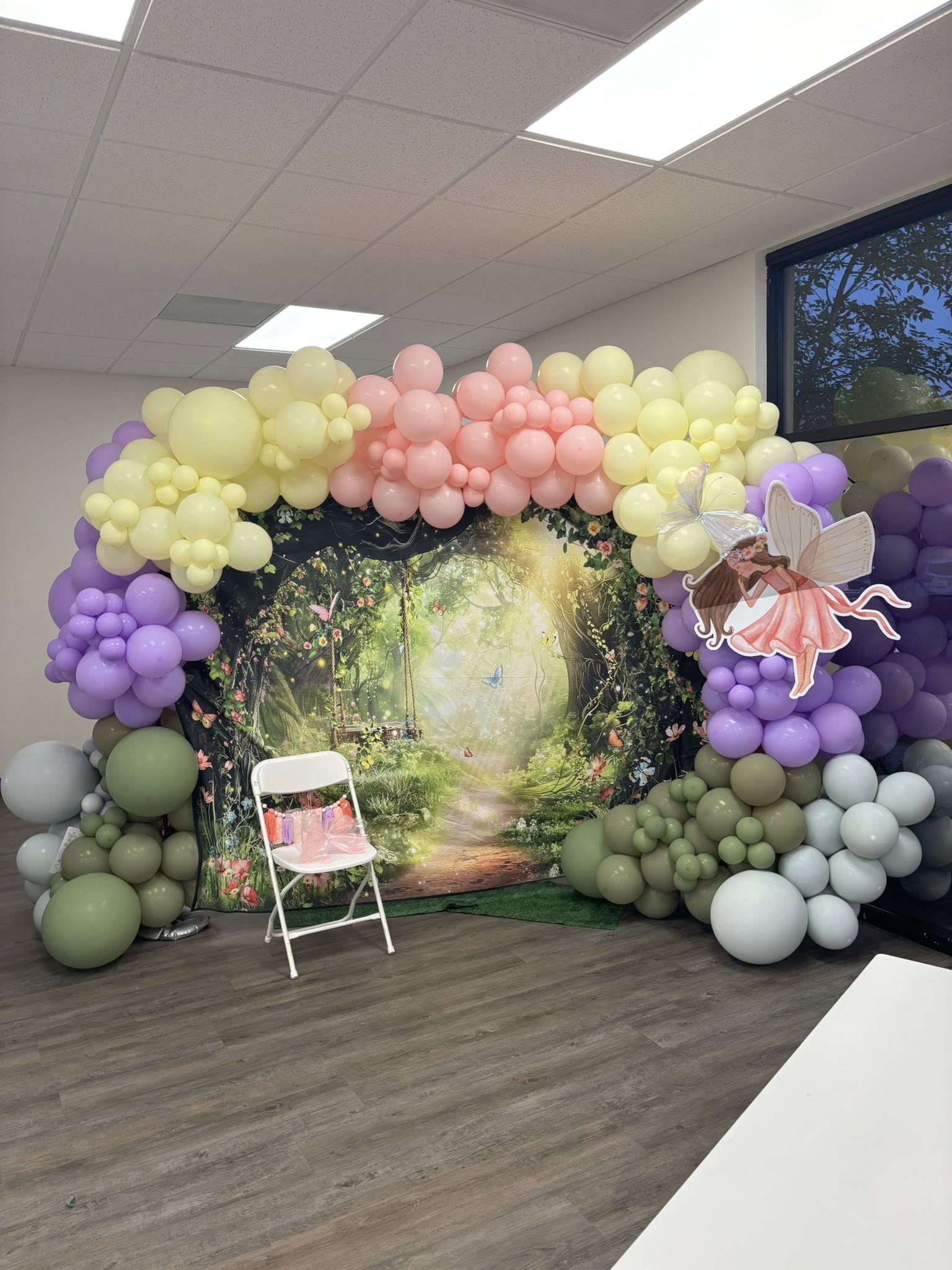 Enchanted forest theme backdrop with a balloon arch, fairy cutout, and chair for party decoration.