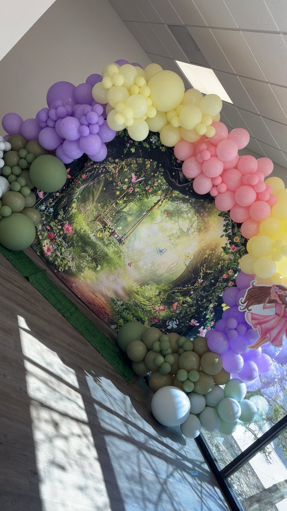 Enchanted forest-themed party backdrop with a fairy cutout and colorful pastel balloon arch.