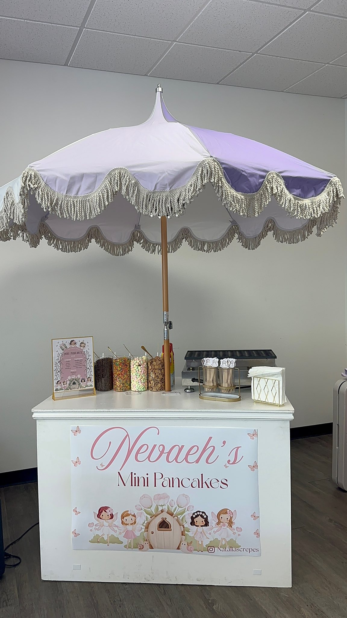Nevaeh's Mini Pancakes catering cart with a lilac fringed umbrella, toppings bar, and pancake griddle.