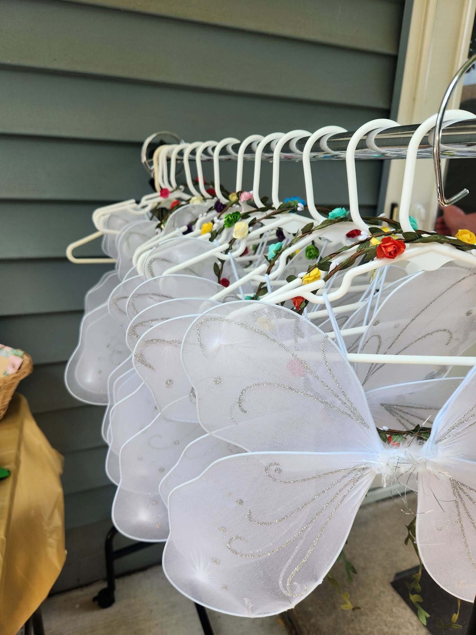 Row of white fairy wings with glitter accents and floral headbands hanging on white coat hangers.