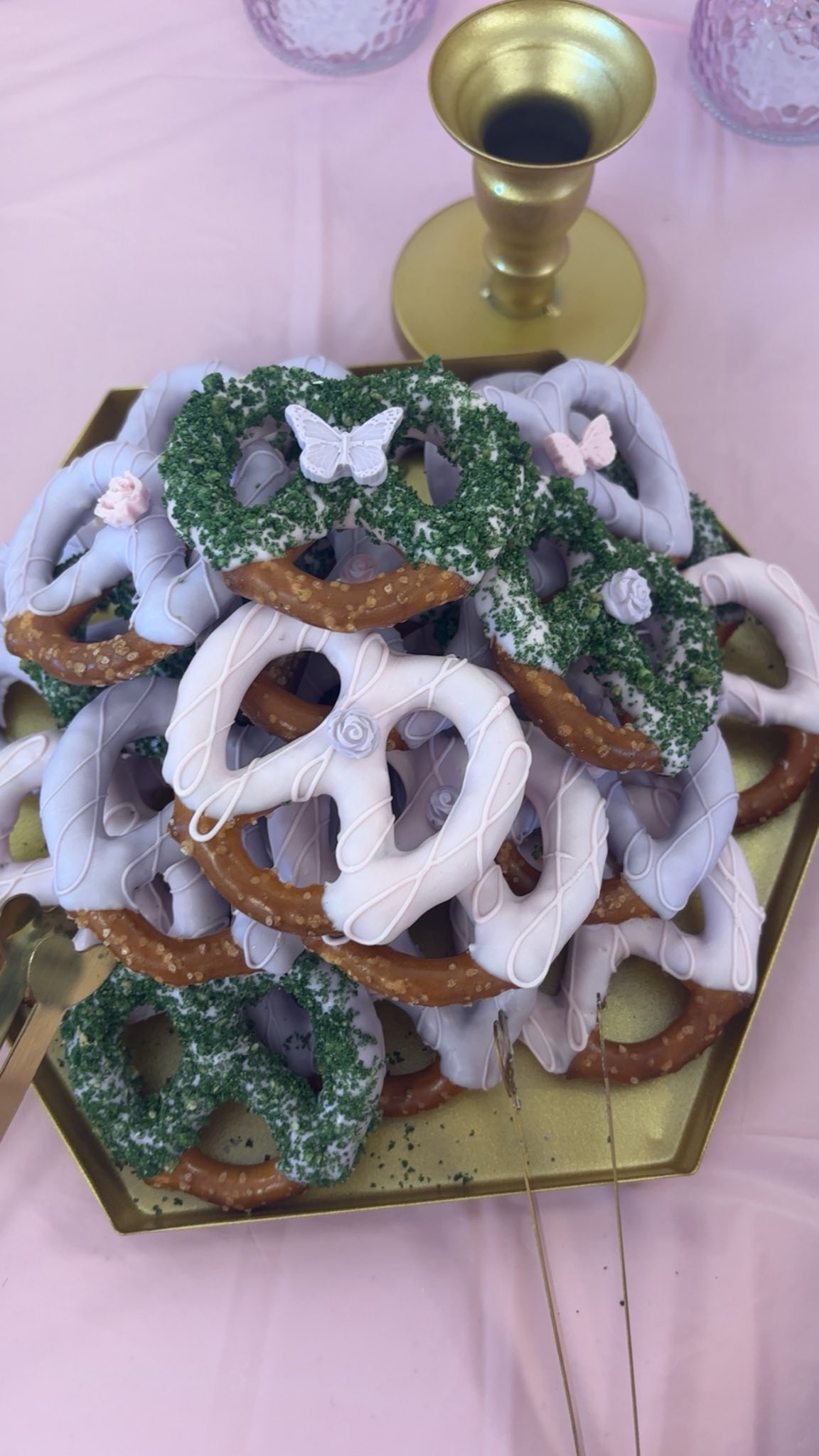 A decorative platter of pretzels dipped in white and light purple icing, decorated with edible green sprinkles, miniature roses, and small butterfly accents.
