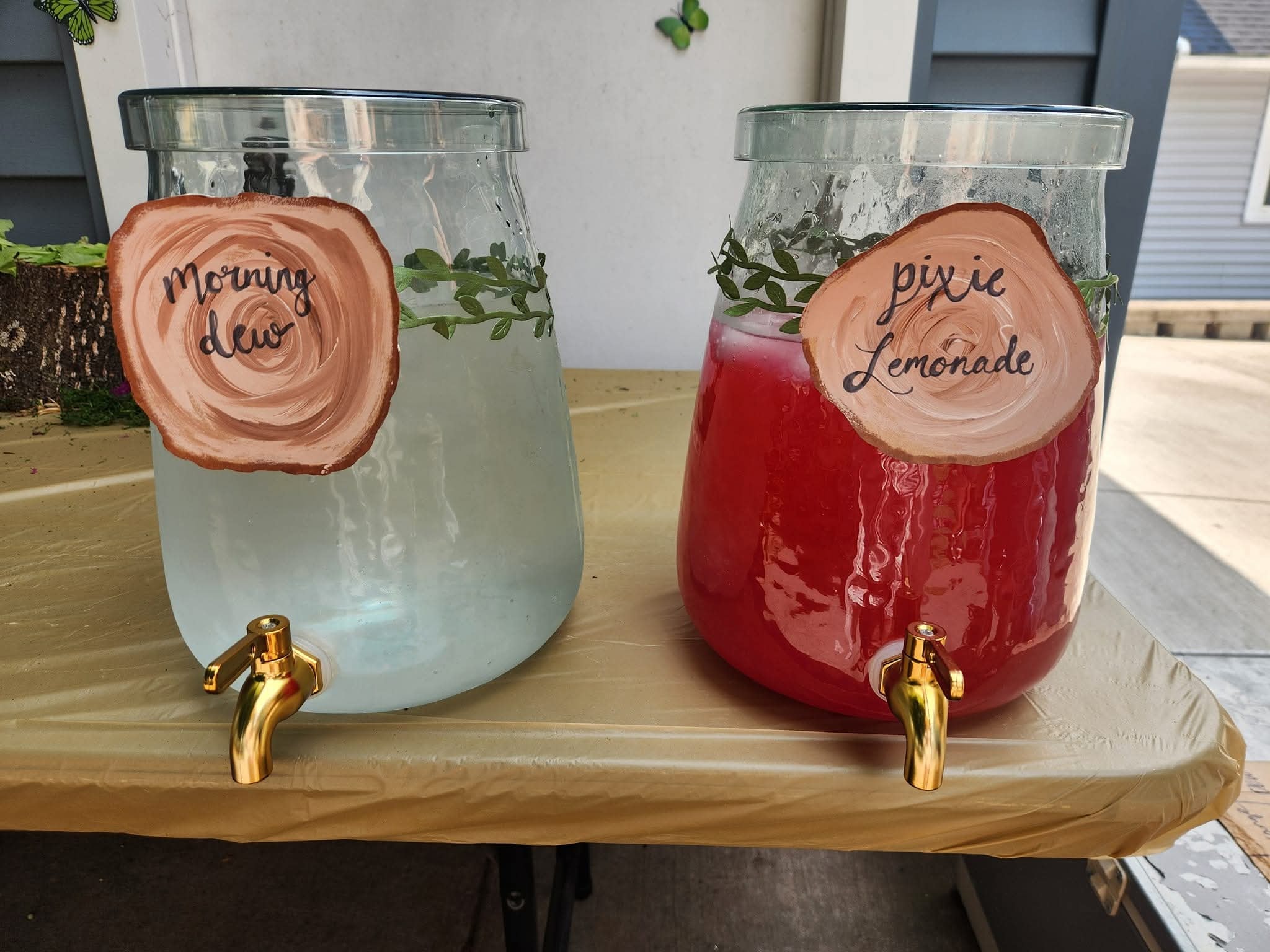 Two glass beverage dispensers labeled Morning Dew and Pixie Lemonade with wood-slice style signs and greenery.