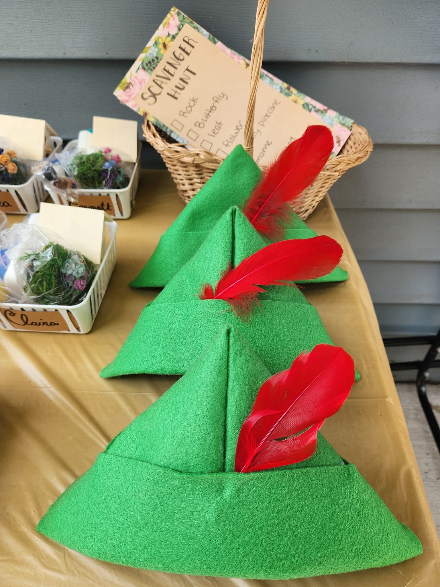 Three green felt Peter Pan-style hats with red feathers, displayed on a table with scavenger hunt supplies.