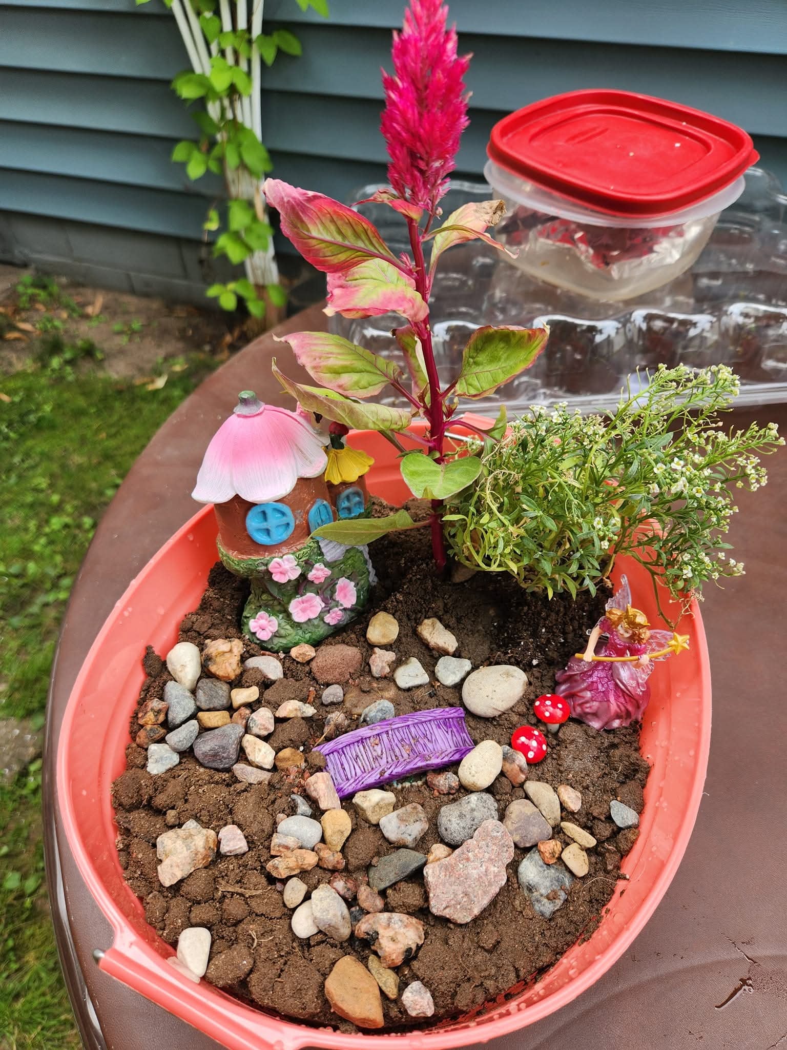 DIY fairy garden in a pink plastic bowl with a toy house, miniature bridge, toadstools, and a fairy figurine.