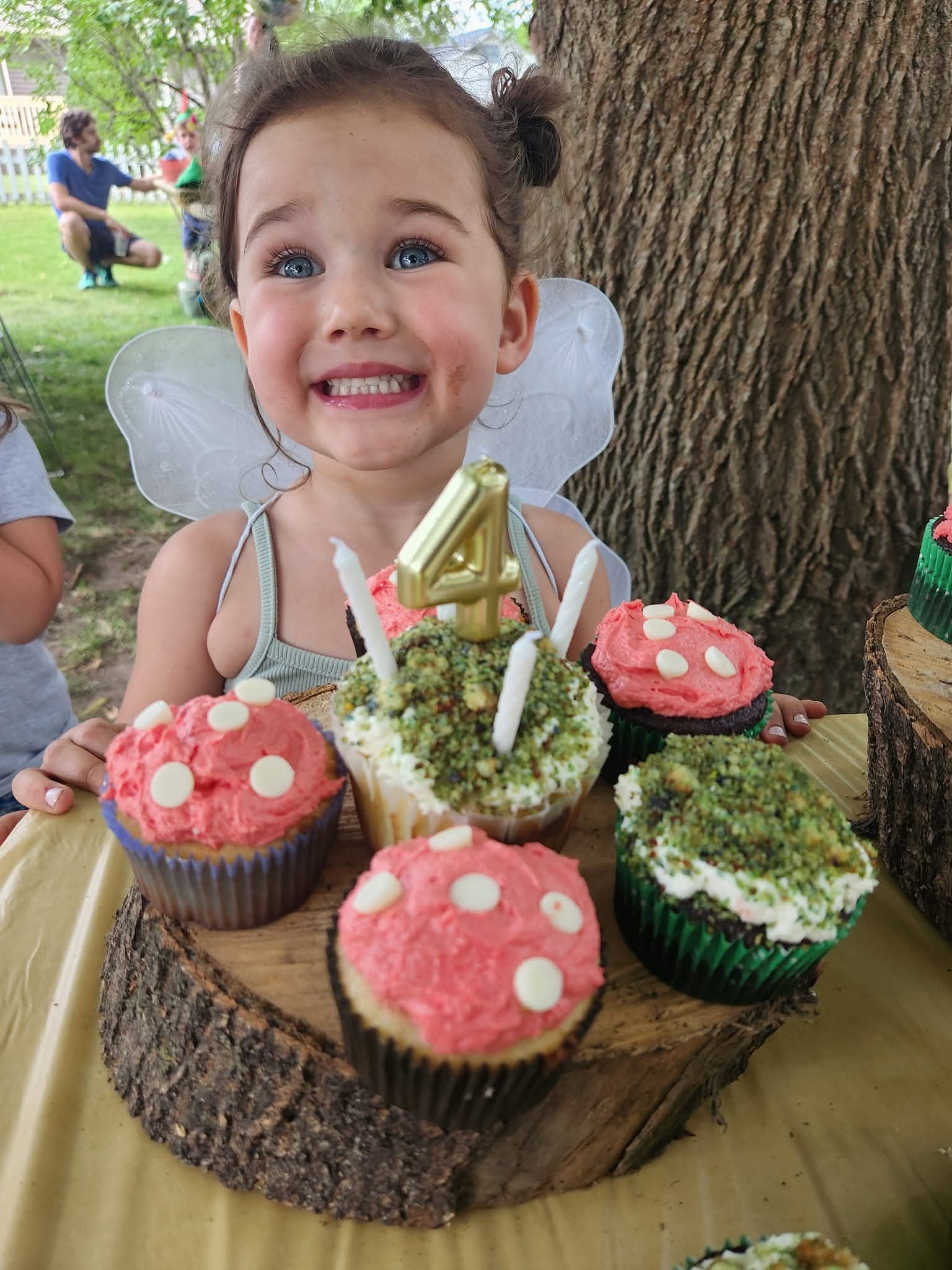 Young girl in fairy wings smiling behind a woodland-themed birthday cupcake display with a number four candle.