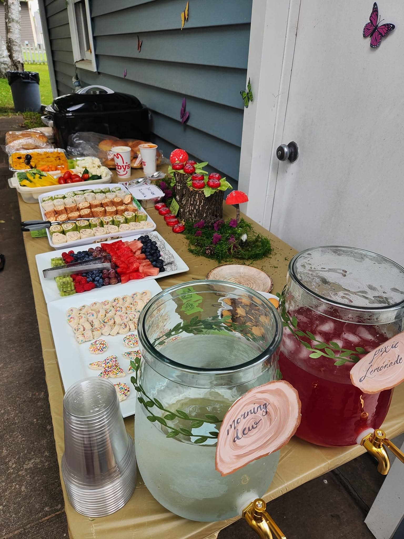 Table set up with party food including drink dispensers labeled Morning Dew to Pixie Lemonade and mushroom decor.