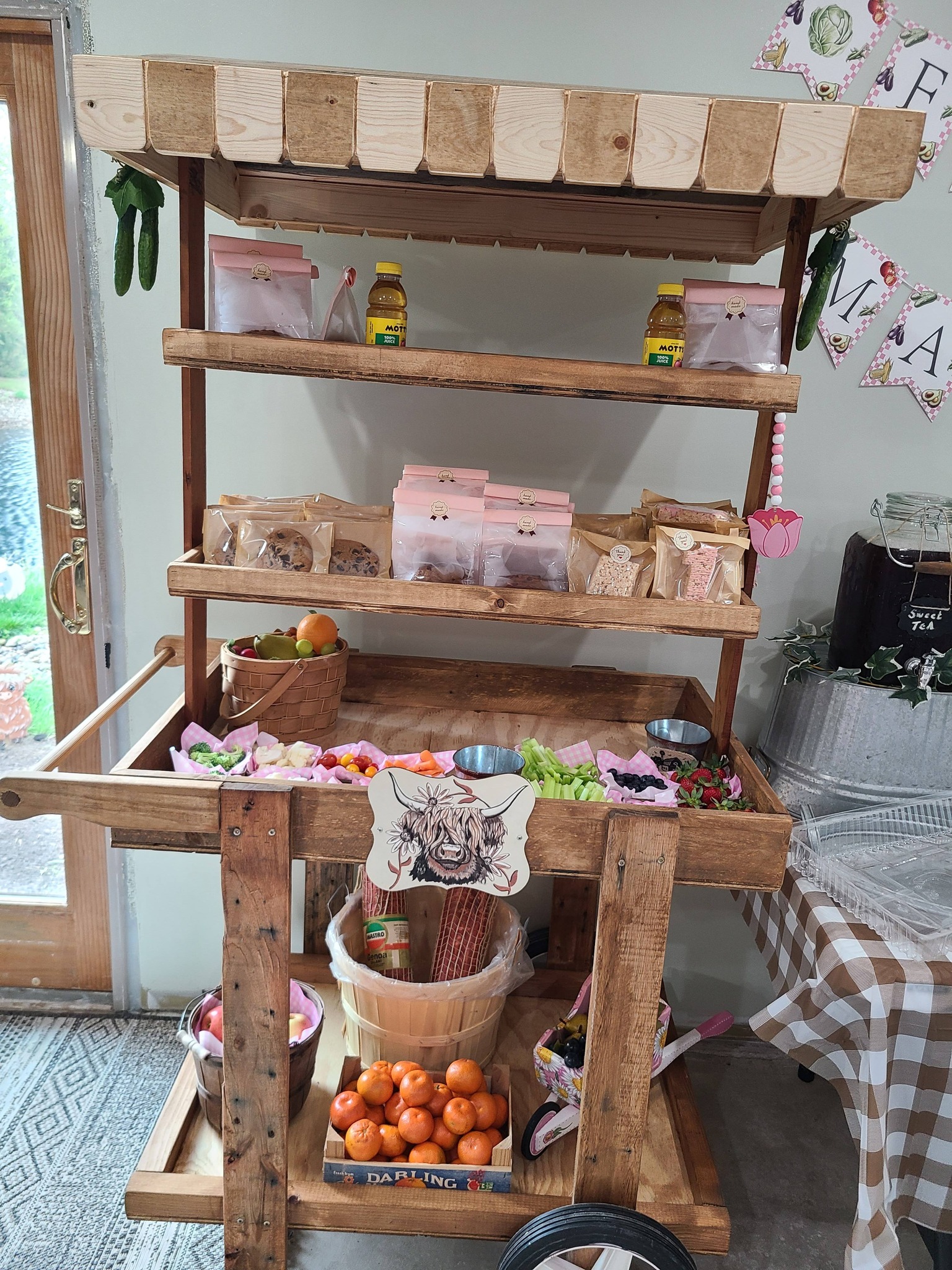 Wooden market-style display cart filled with snacks, fresh fruits, vegetables, and pre-packaged treats at a party.