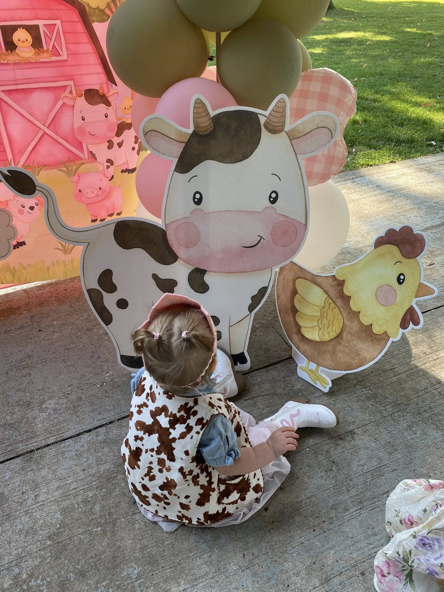 Toddler in a cow-print vest sitting outside in front of farm-themed party decorations including cow and chicken cutouts.