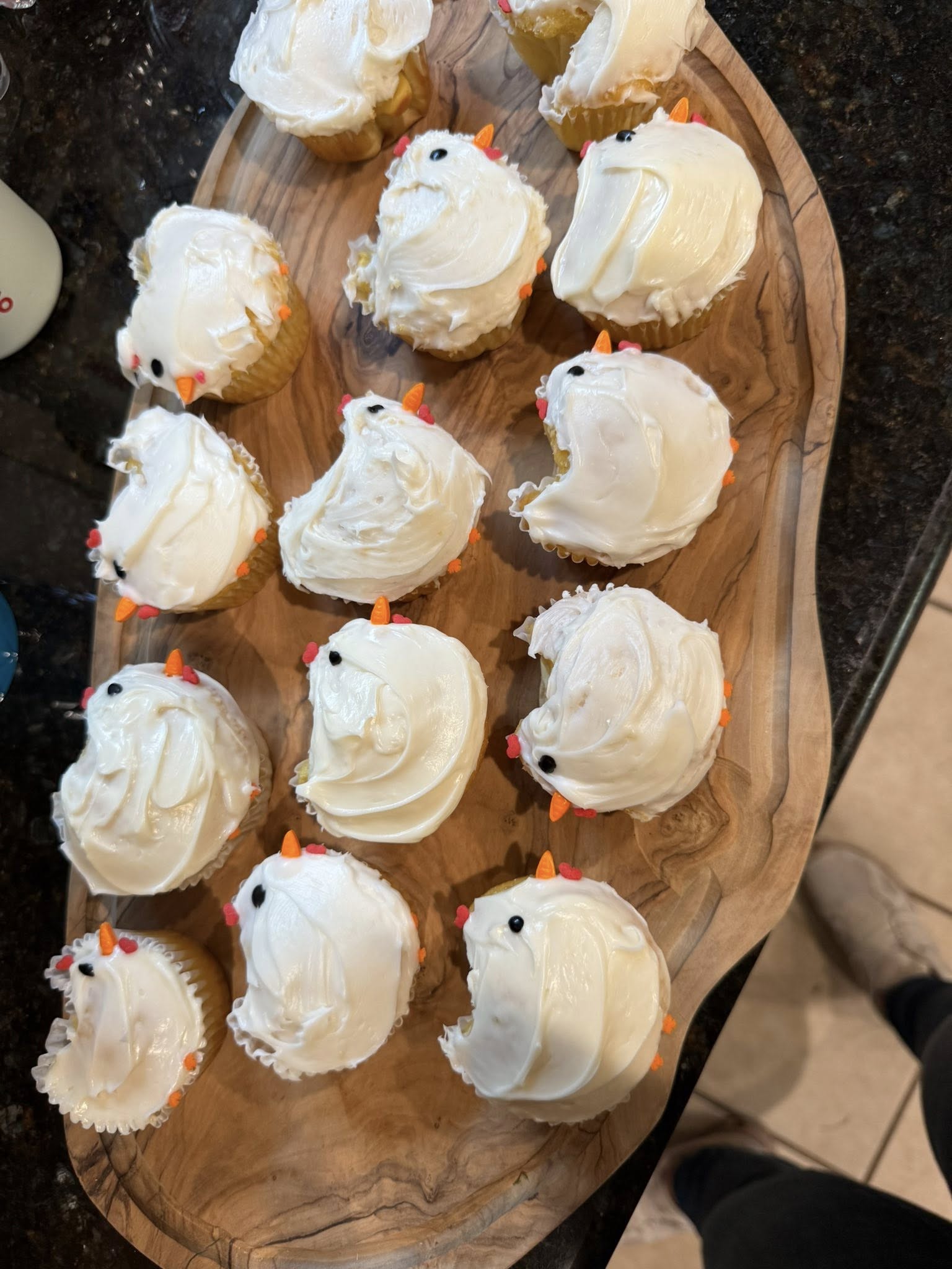 Homemade cupcakes decorated to look like little chicks with white frosting, orange beaks, and black eyes on a wooden board.