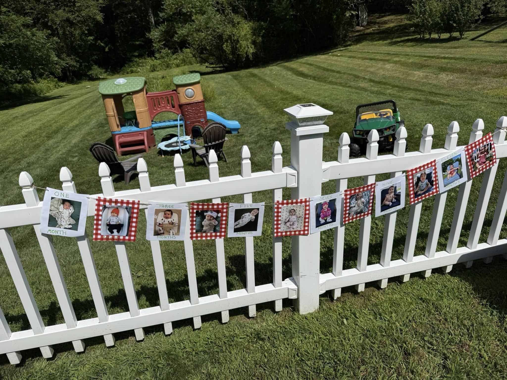 Photo banner featuring a baby's monthly milestones hung on a white picket fence in a backyard.
