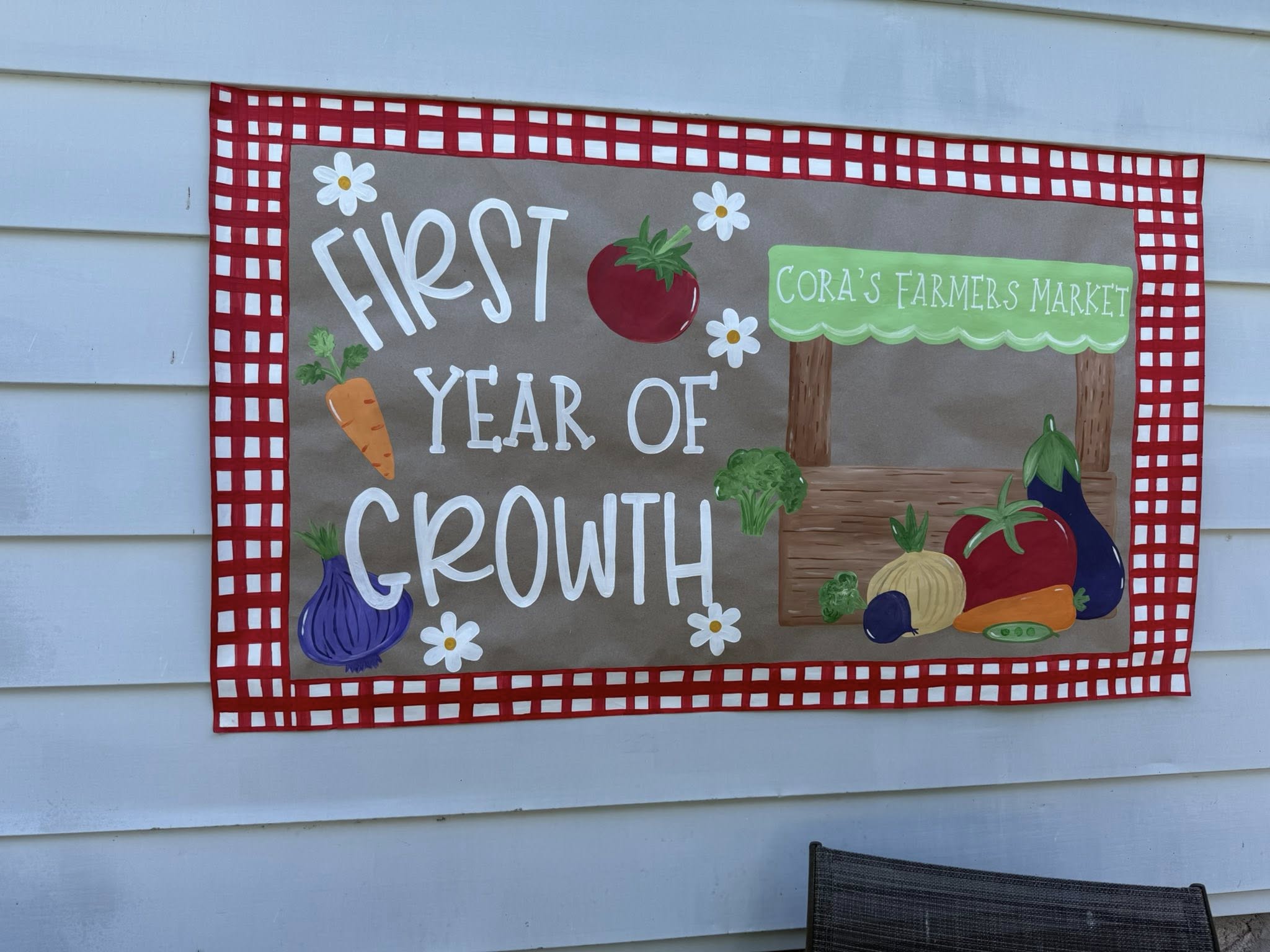 A decorative sign reading Cora's Farmers Market First Year of Growth, featuring illustrations of vegetables.