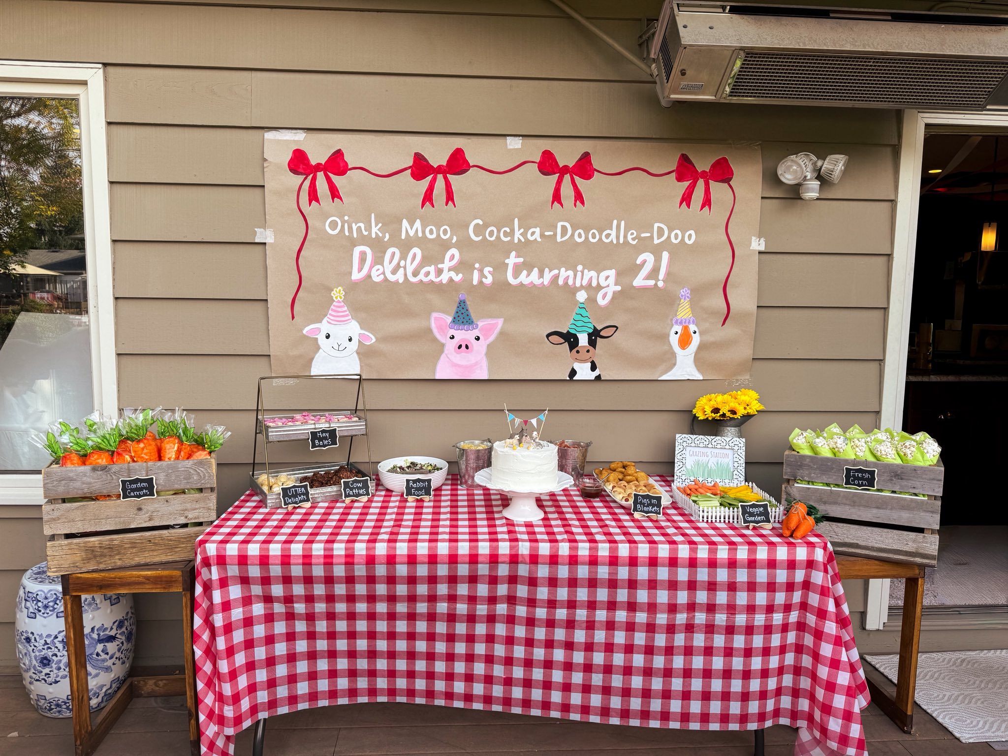 A party table decorated for a second birthday with a red-and-white checkered tablecloth, farm-themed snacks, and a banner that reads Oink, Moo, Cocka-Doodle-Doo, Delilah is turning 2.