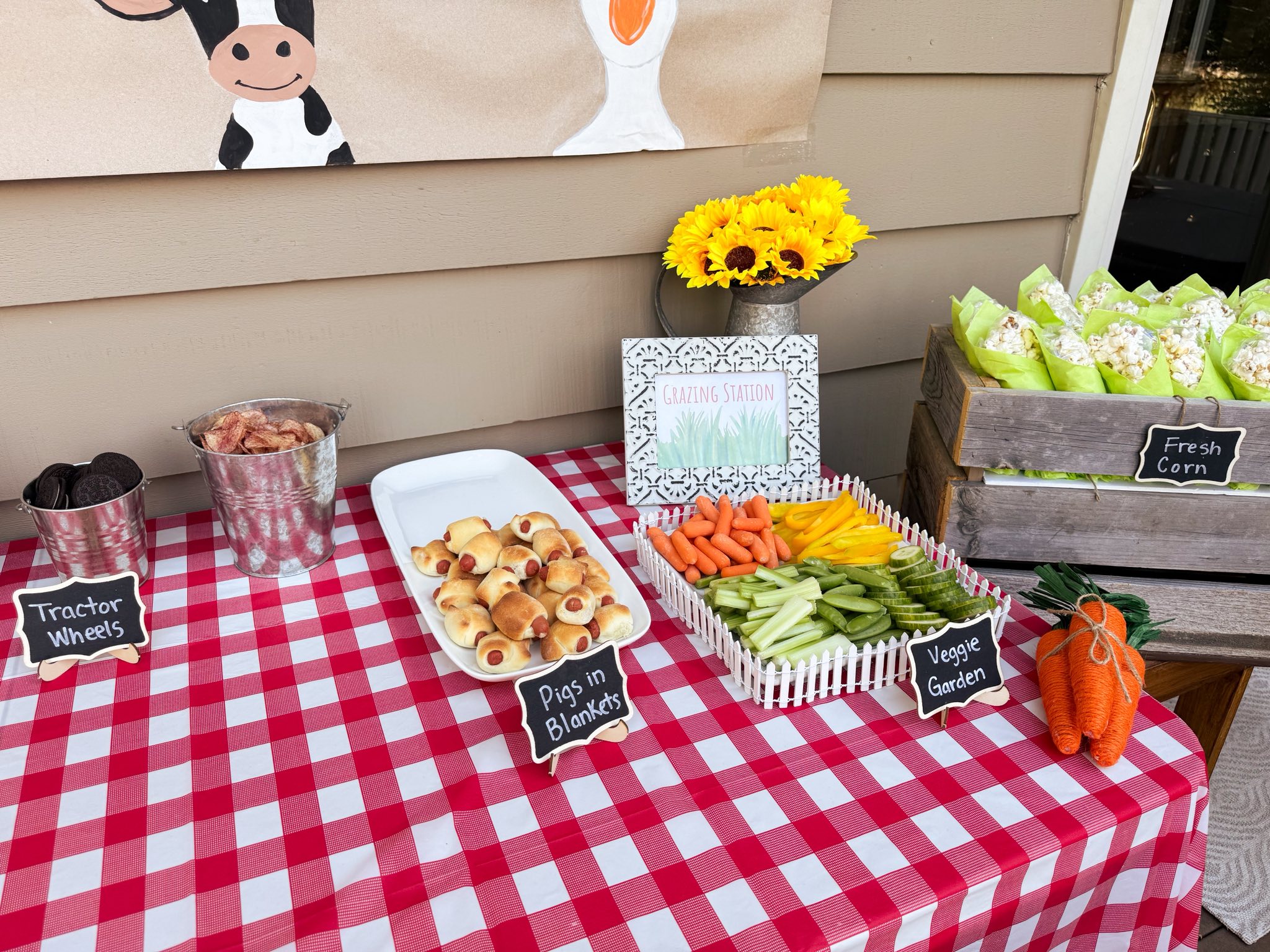 Grazing station party table with labeled snacks like pigs in blankets, veggie garden, and tractor wheels.
