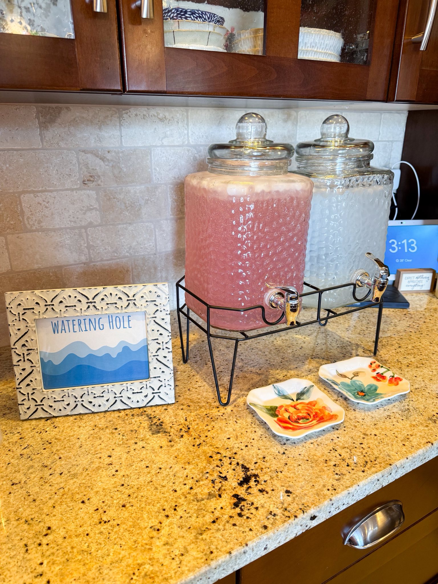 Two glass beverage dispensers on a metal stand next to a decorative Watering Hole sign on a kitchen counter.