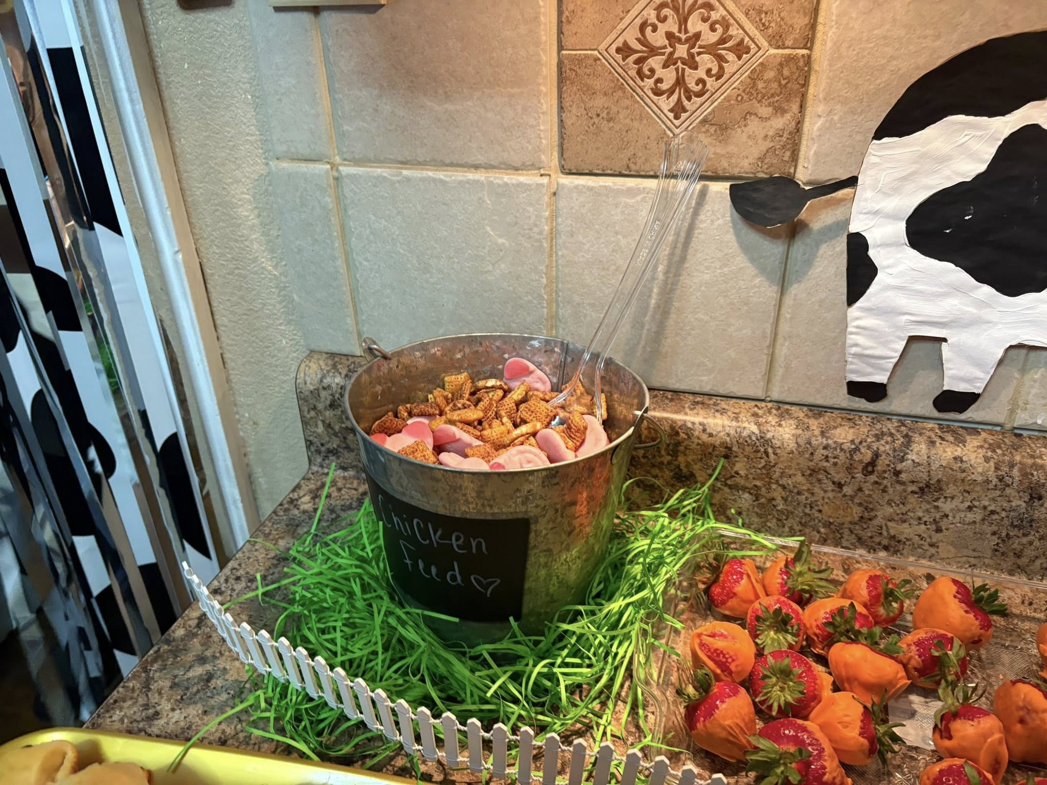 Metal bucket labeled Chicken Feed filled with snack mix and gummy farm animal treats on a buffet table.