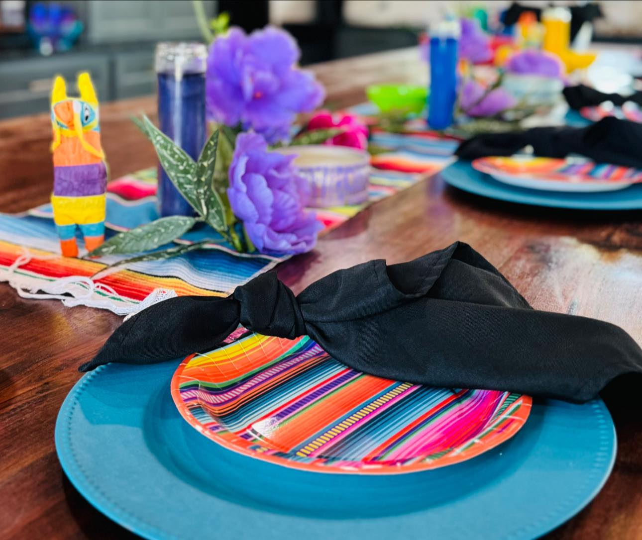 Fiesta themed table setting with colorful striped plates, teal chargers, black napkins, and a mini piñata centerpiece.