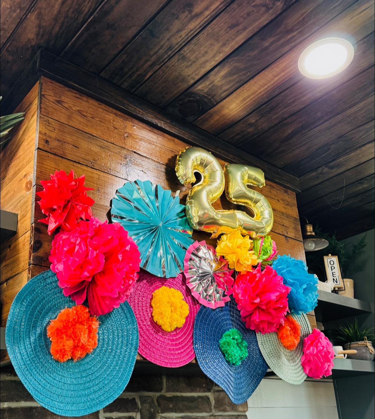 Festive wall decoration with gold number 25 balloons, colorful paper fans, and vibrant tissue paper pom-poms.