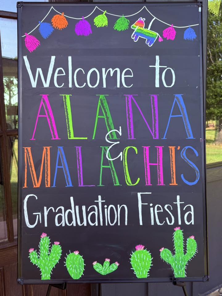 Blackboard welcome sign for Alana and Malachi's graduation fiesta decorated with colorful pom-poms and cacti.