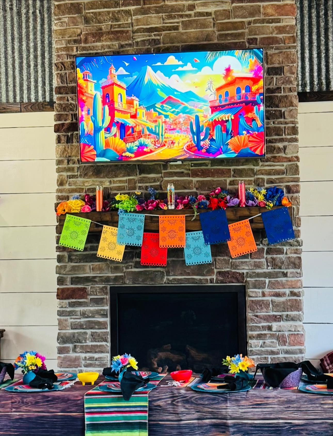 Fiesta themed fireplace mantel decorated with colorful papel picado bunting, bright floral arrangements, and a vibrant landscape display on the mounted TV.