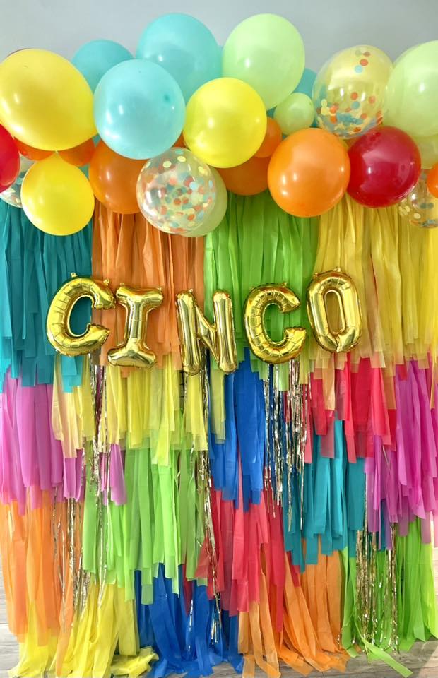 Cinco de Mayo party decoration with colorful balloons and a 'CINCO' banner against a multi-colored fringe backdrop.
