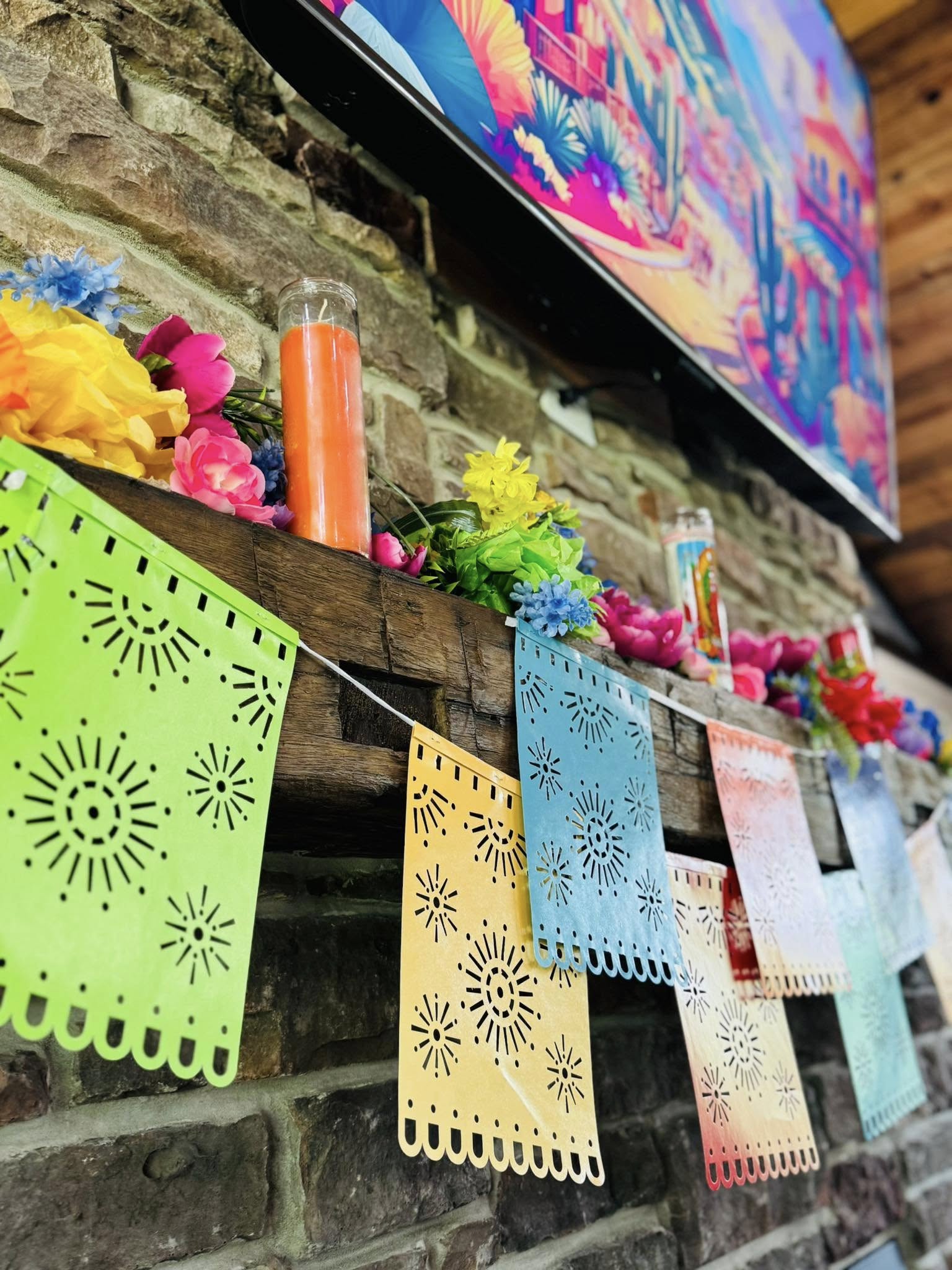 Colorful papel picado banner hanging from a rustic stone fireplace mantle decorated with vibrant paper flowers.