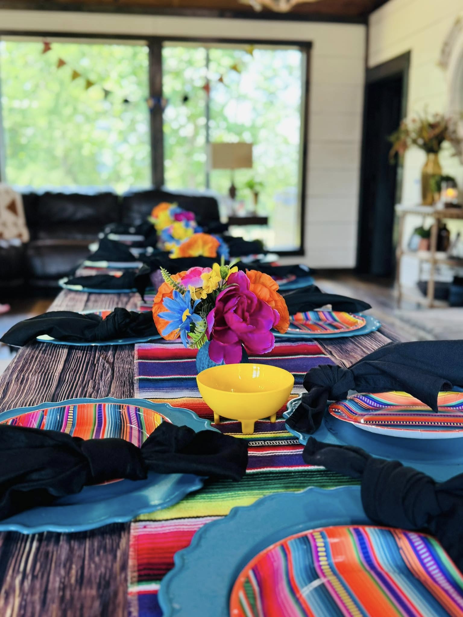 Dining table set with colorful striped table runner, blue plates, black napkins, and bright floral centerpieces.