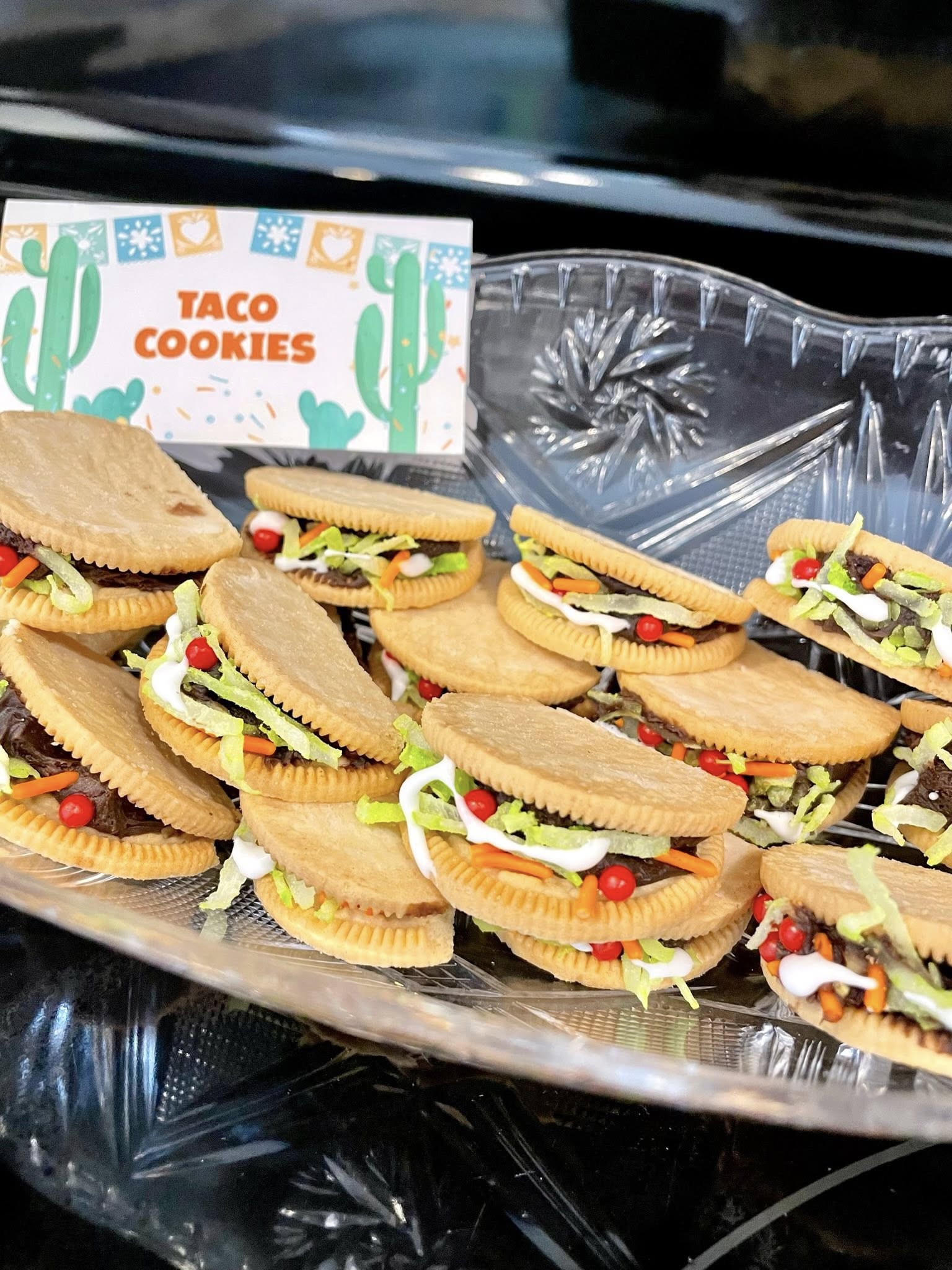 Platter of taco-shaped sandwich cookies decorated with shredded coconut lettuce and red icing tomatoes.
