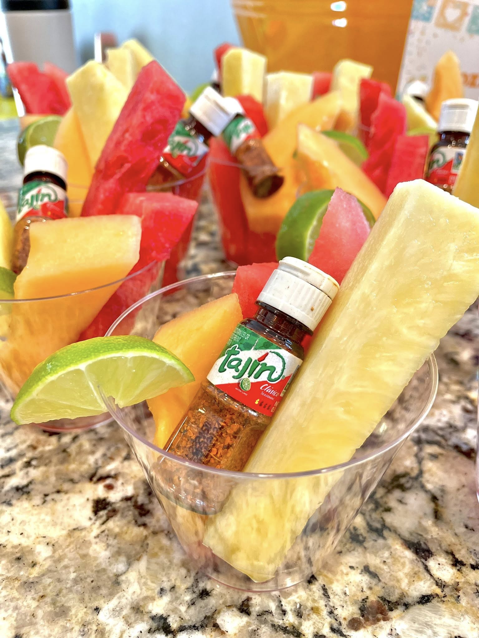 Clear plastic cups filled with fresh pineapple, watermelon, and cantaloupe slices, garnished with a lime wedge and a mini bottle of Tajín seasoning.