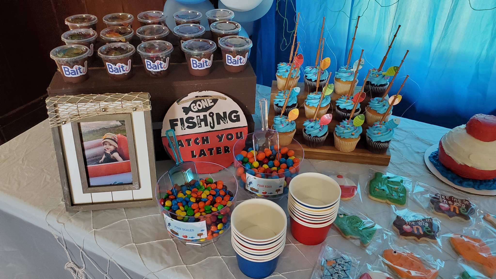 Fishing-themed first birthday party dessert table with cupcakes, candy 'fish scales', pudding cups, and fish cookies.