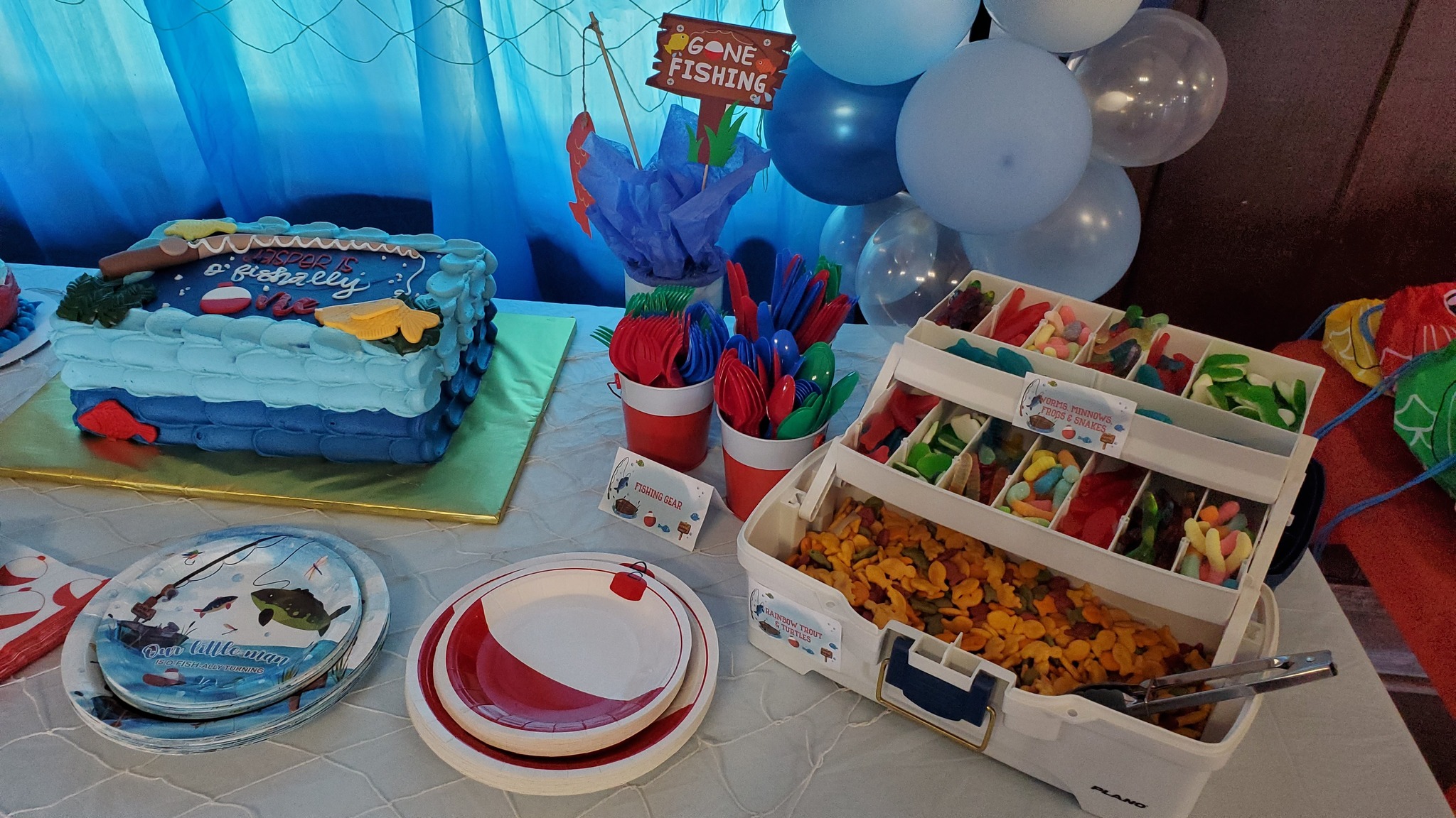 Fishing-themed first birthday party table with a decorated cake, gummy candy snacks in a tackle box, and fishing-themed plates, napkins, and cutlery.