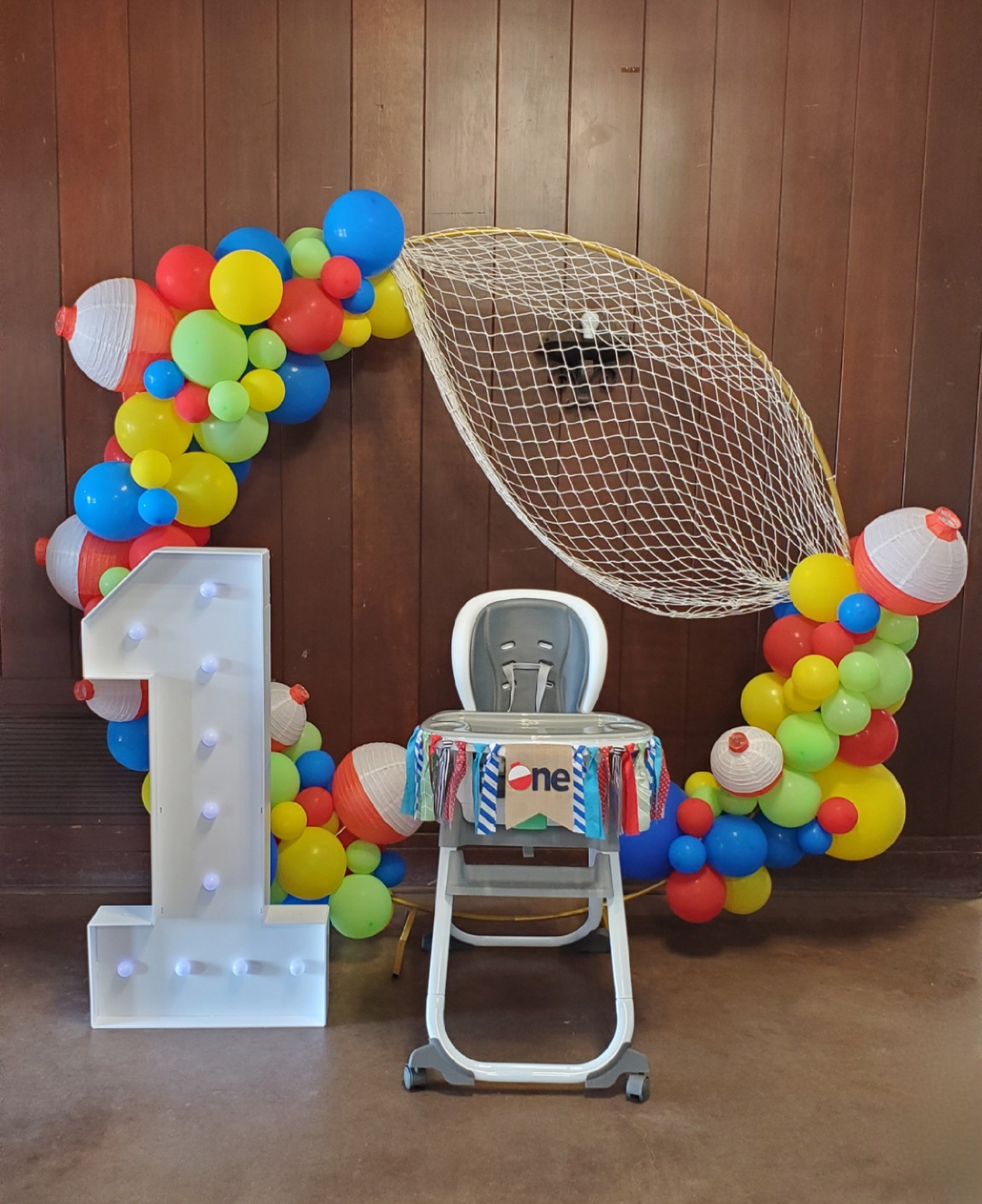 Fishing-themed first birthday party setup with a balloon garland, fishing net, light-up number one, and high chair