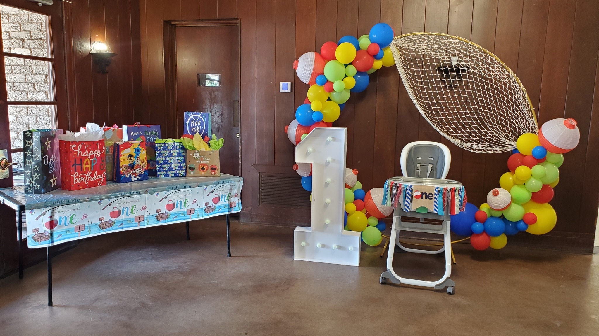 First birthday party setup featuring a fishing-themed balloon garland, gift table, and a high chair with a one sign