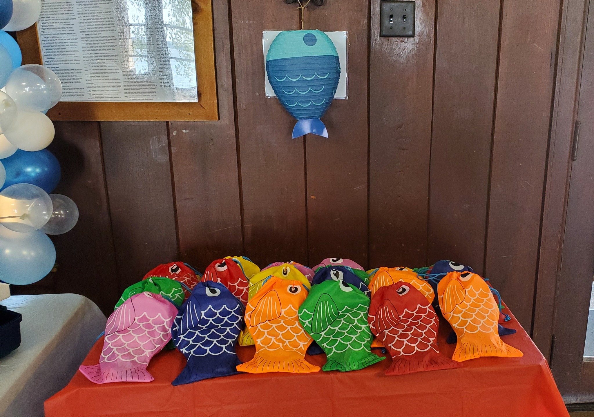 Colorful fish-shaped drawstring gift bags arranged on a red tablecloth with a blue fish lantern wall decor.