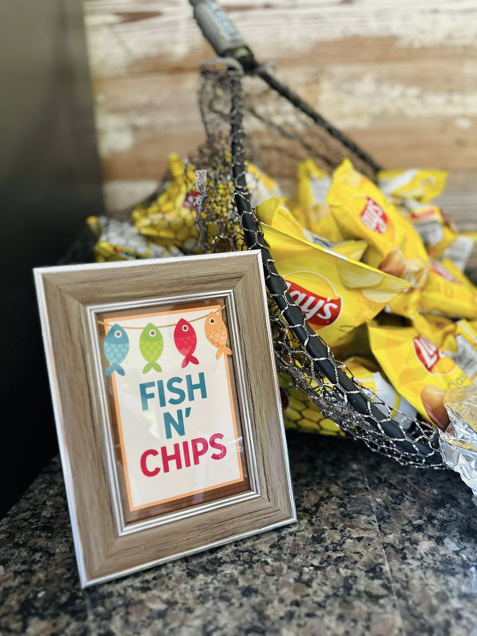 Table sign labeled Fish N' Chips placed next to a fishing net filled with bags of Lay's potato chips.