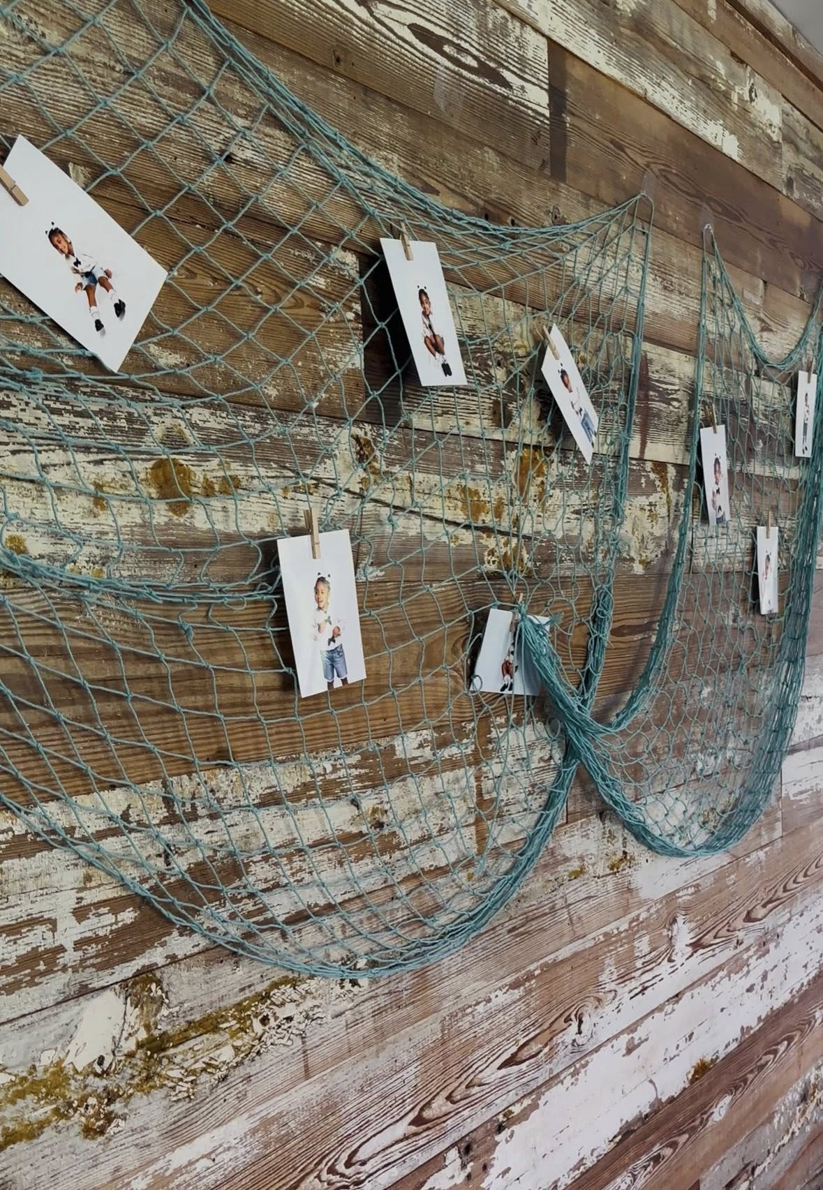 Teal decorative fishing net wall display hanging photos with wooden clothespins against a weathered wood wall.