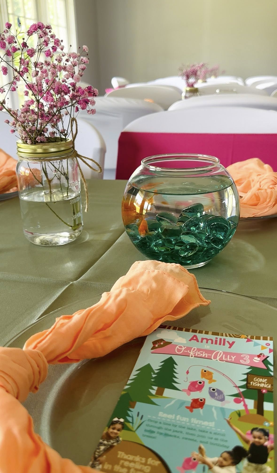 Decorated event table with a goldfish bowl centerpiece, pink baby's breath in a mason jar, and fishing themed invitation