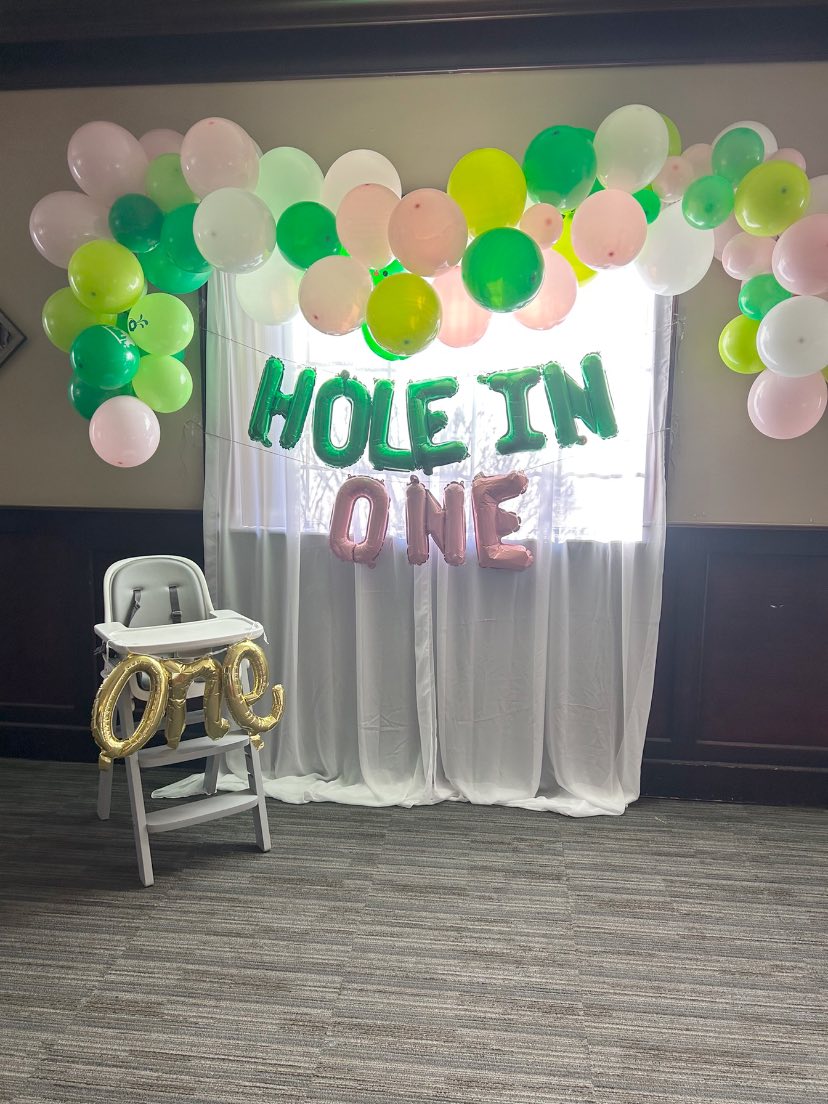 Hole in One birthday party decoration setup with a high chair, balloon arch, and hanging letter balloons.