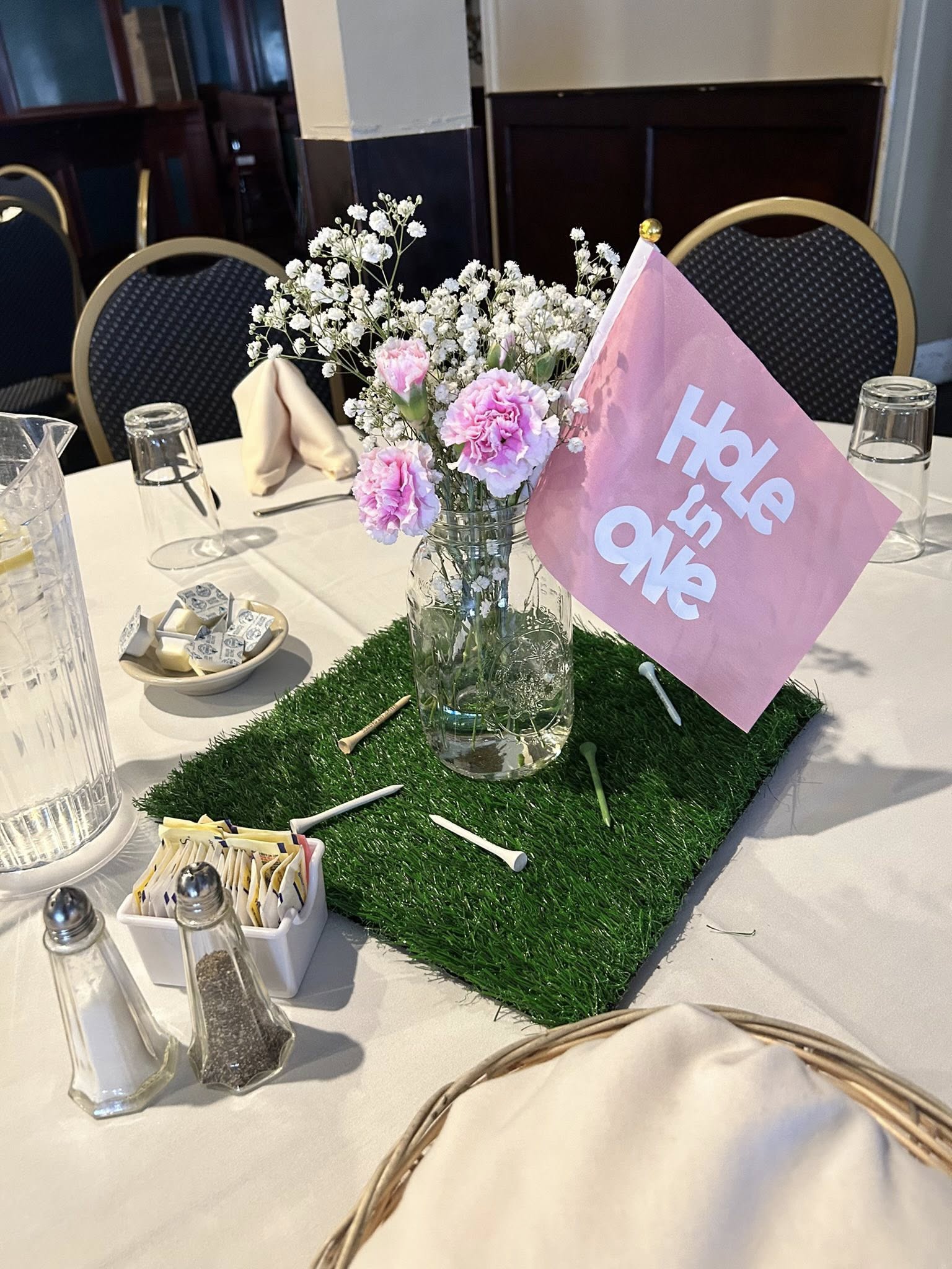Table centerpiece featuring a mason jar with pink carnations and baby's breath, a pink 'Hole in One' flag, and golf tees on an artificial grass mat.