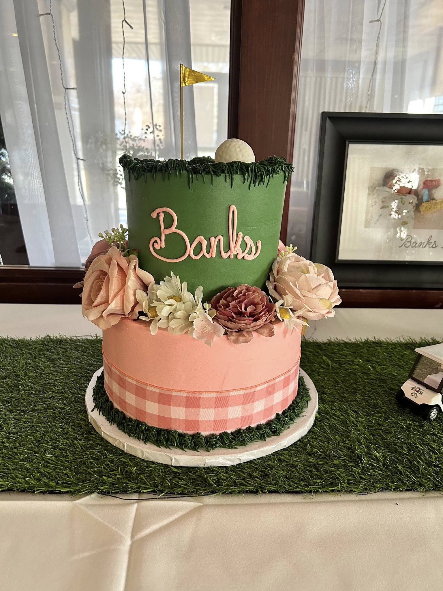 Two-tier golf-themed cake with green grass frosting, a golf ball and flag topper, and pink floral accents.