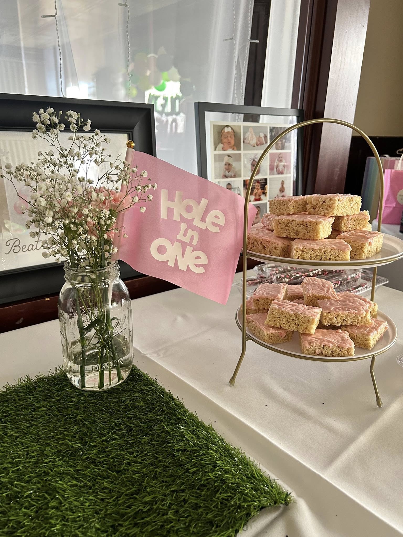 Two-tier stand filled with pink-glazed rice crispy treats next to a baby shower centerpiece with flowers and a Hole in One flag.