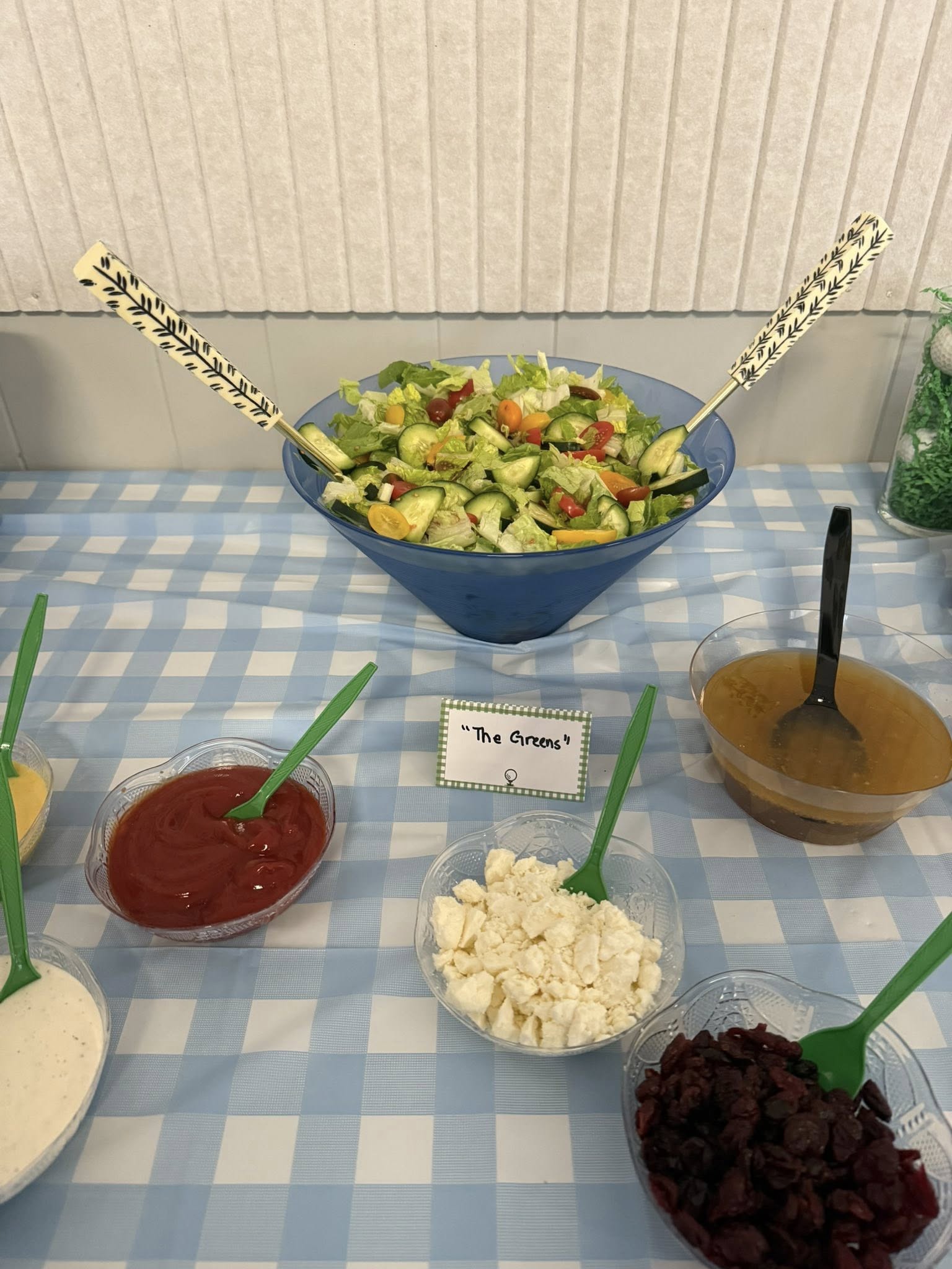 Large blue bowl of green salad with cucumber and cherry tomatoes, accompanied by various bowls of dressings and toppings.