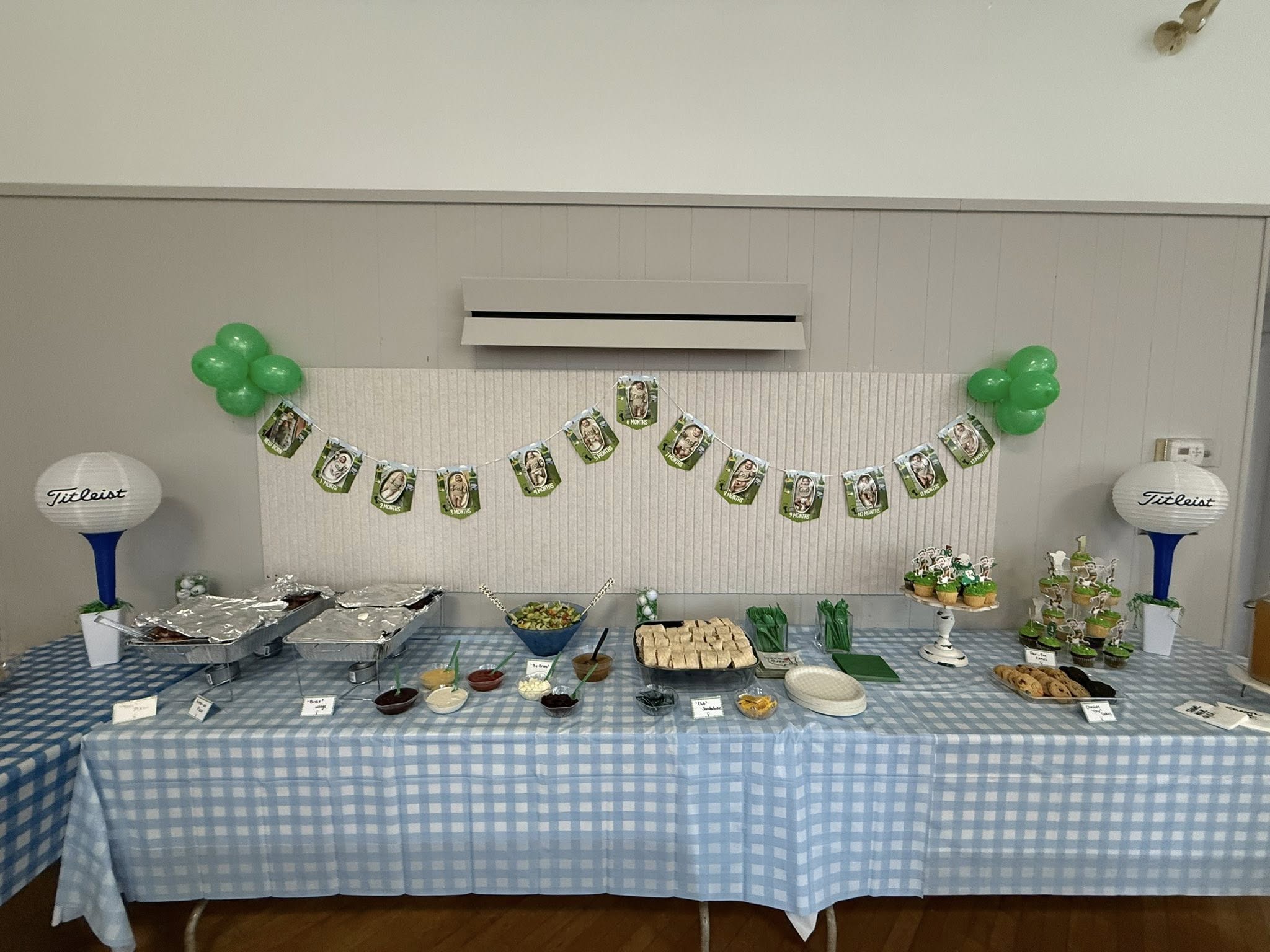 Golf-themed birthday party buffet table with a banner showing baby monthly photos, plaid tablecloths, and food.