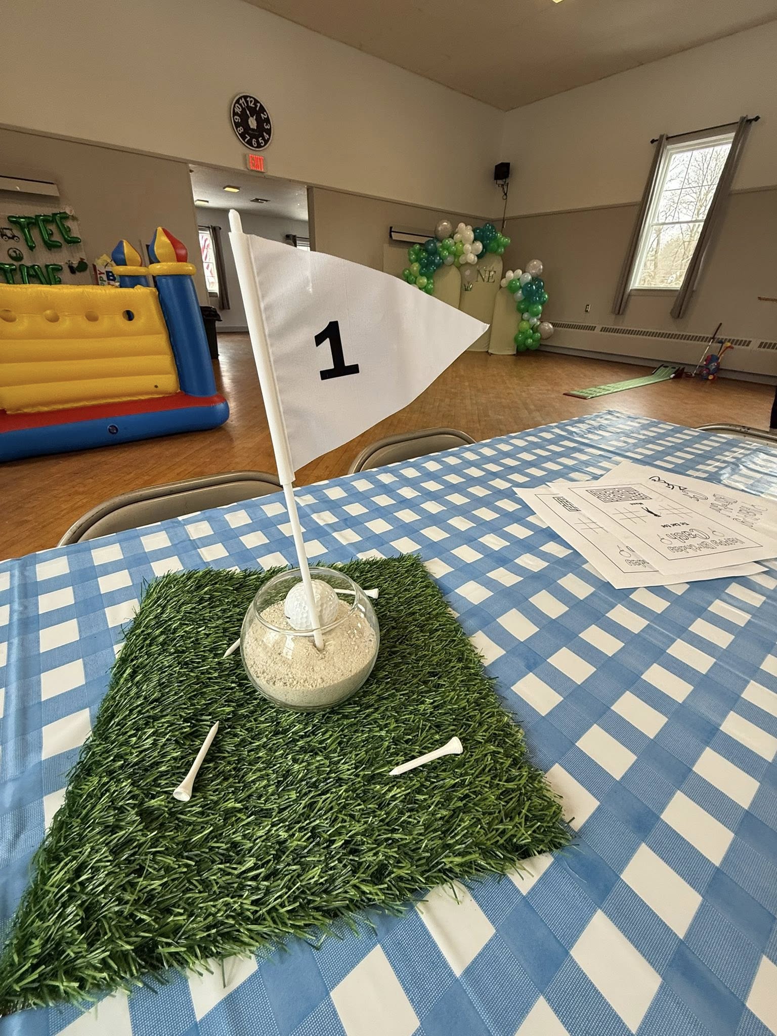 Golf-themed table centerpiece with artificial grass mat, sand, golf ball, and a number 1 flag on a blue checkered tablecloth.