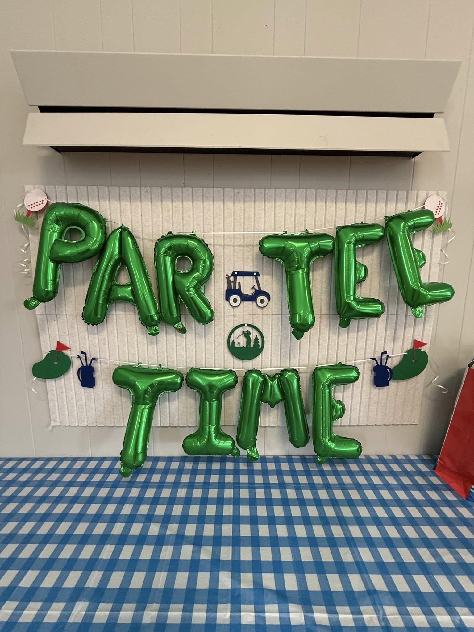 Green balloon banner on a wall spelling Par-Tee Time with golf-themed decorations above a blue checkered table.